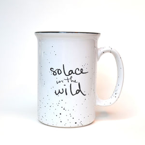 White ceramic Solace in the Wild mug with black writing and black speckles.